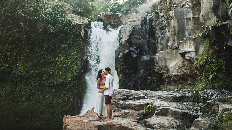 Young Couple Kissing With View Waterfall. Happy Together, Honeymoon In Bali