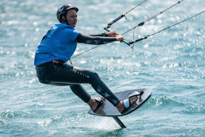 Woman in wetsuit, helmet stretching out riding kitefoil board, hanging onto sail bar
