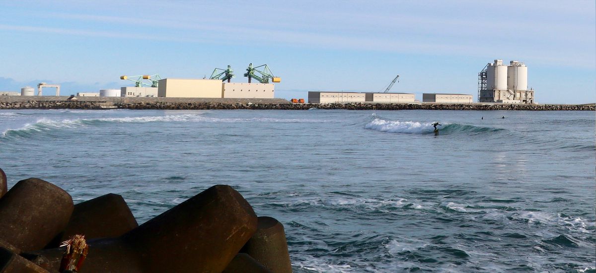 A photo from Kitaizumi Beach focused on a man in the distance surfing a wave. In the background are industrial buildings.