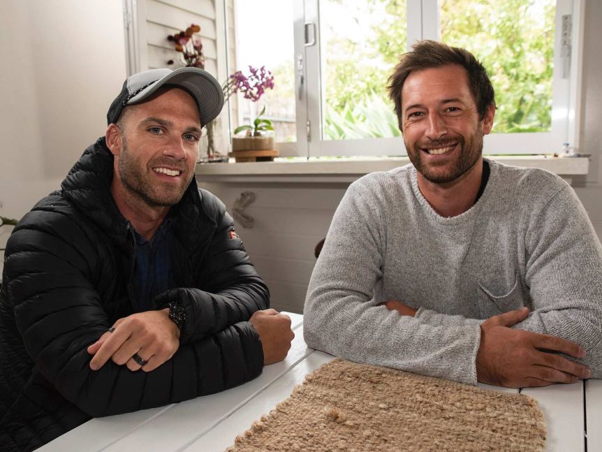 Two men sit at an indoor table and smile at the camera.