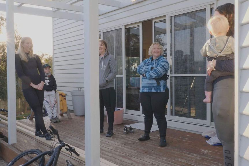 A group of ladies in wetsuits and children, on a porch before surf