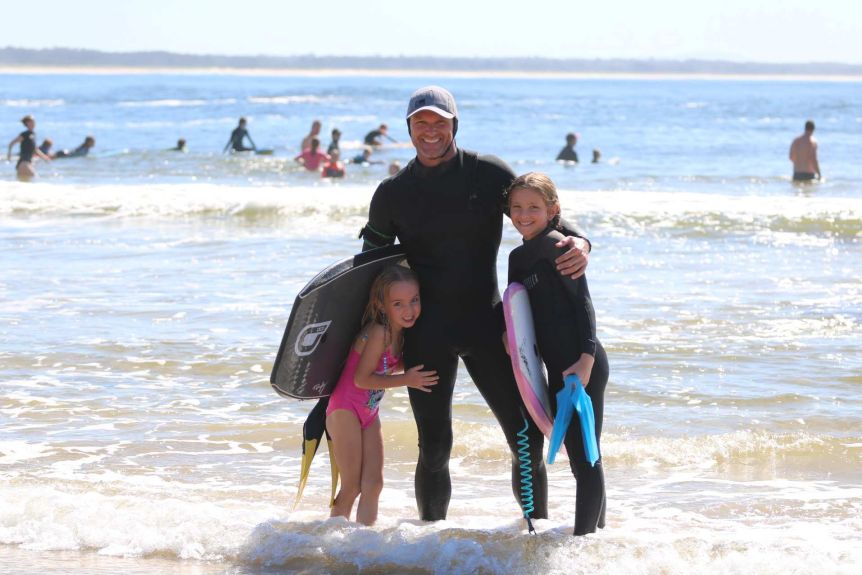 A man in a full wetsuit stands in the surf with his arms around his two young daughters.