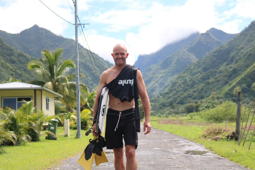 A man in board shorts walks smiling down a dirt road on a tropical island carrying flippers and a bodyboard.