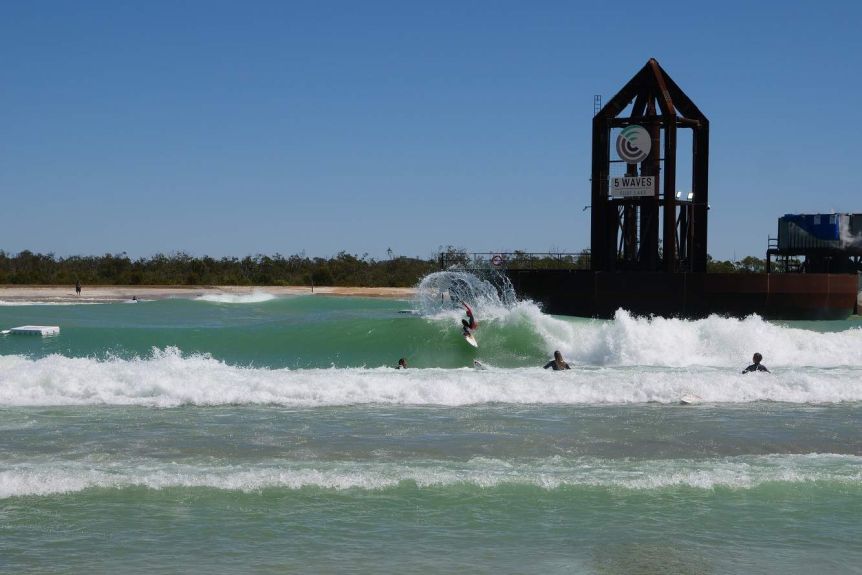 Waves crash into the shore of a wave pool, a woman surfs on one of waves sending spray off the back of the wave.