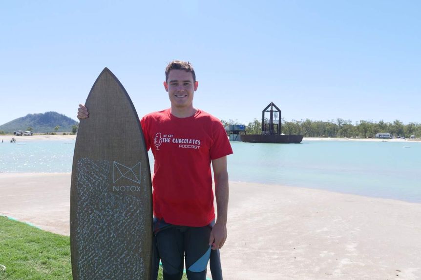 A man stands in a wetsuit holding a surfboard on the shore of a wave pool.